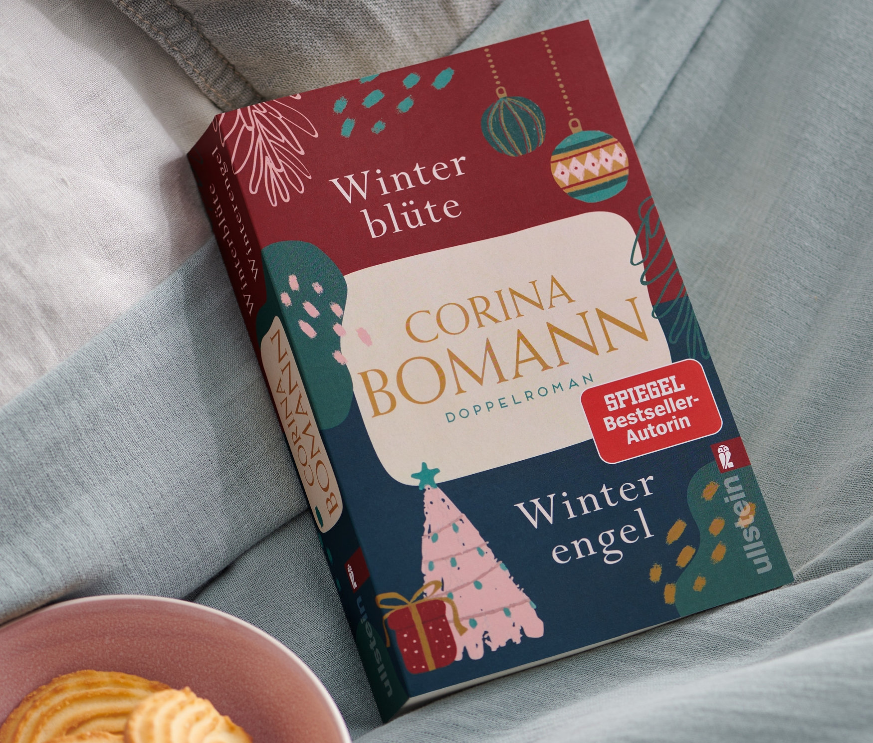 Auf hellblauem Stoff liegt das Buch "Doppelroman: »Winterblüte« und »Winterengel«", daneben eine rosa Schale mit Keksen.