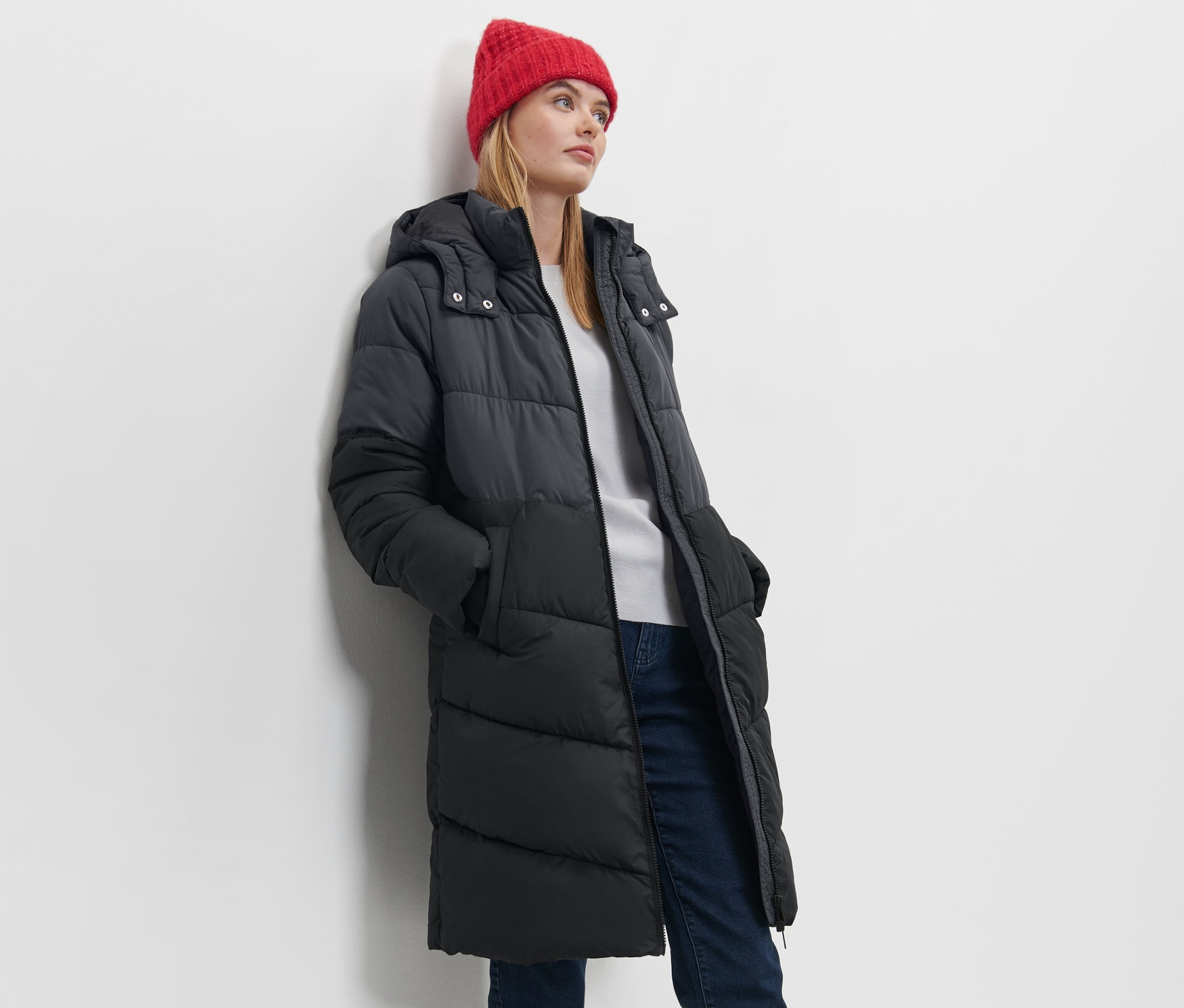 Frau posiert in Steppmantel mit Kapuze und roter Mütze.
