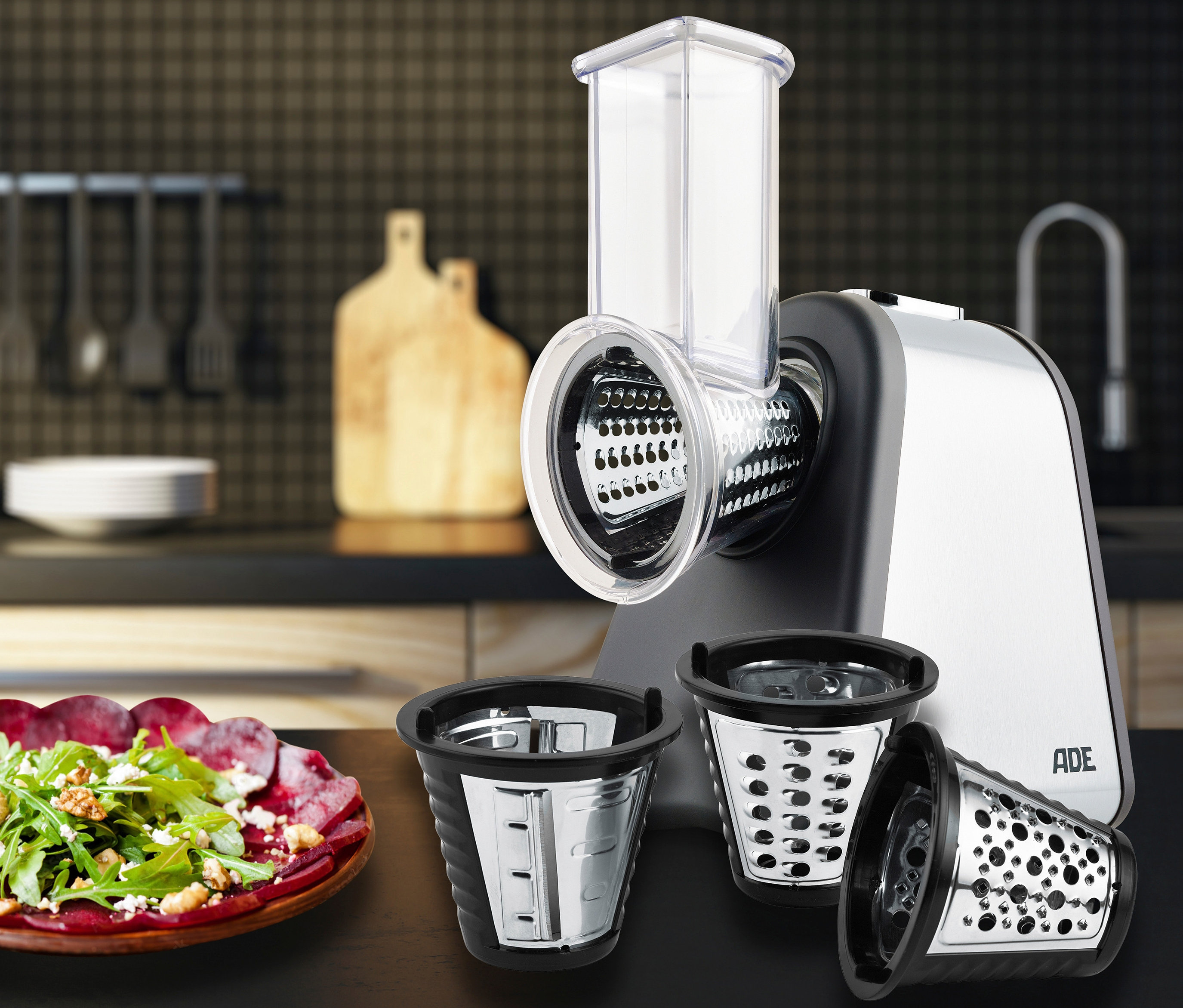 ADE Elektrische Reibe »KA1802« mit drei Aufsätzen auf einem Tisch neben einem Salatteller.