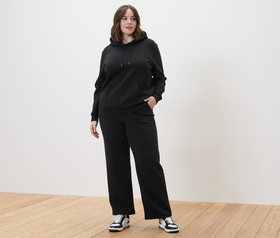 Frau in schwarzem Hoodie und einer Sweathose steht auf einem Holzboden.