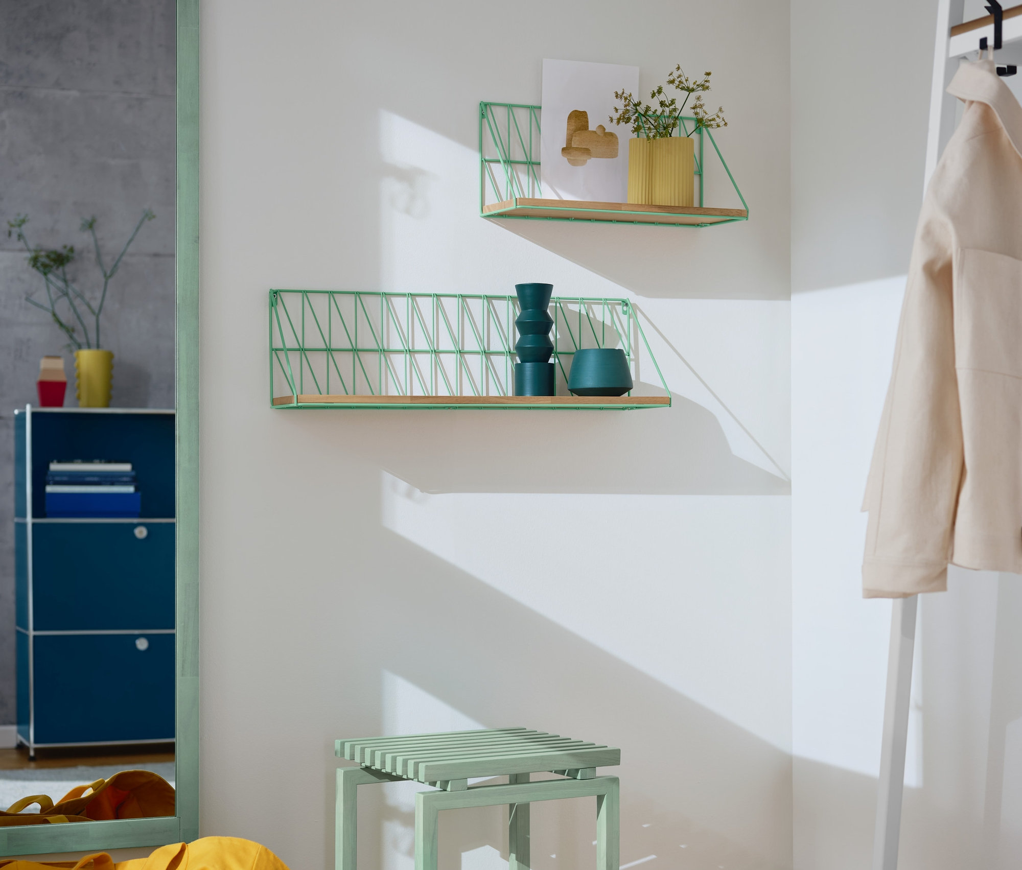 Eckansicht eines Zimmers mit einem Ganzkörperspiegel mit Rahmen in Salbei, zwei Wandregalen in Salbei, einem salbeifarbenen Hocker und einer gelben Tasche.