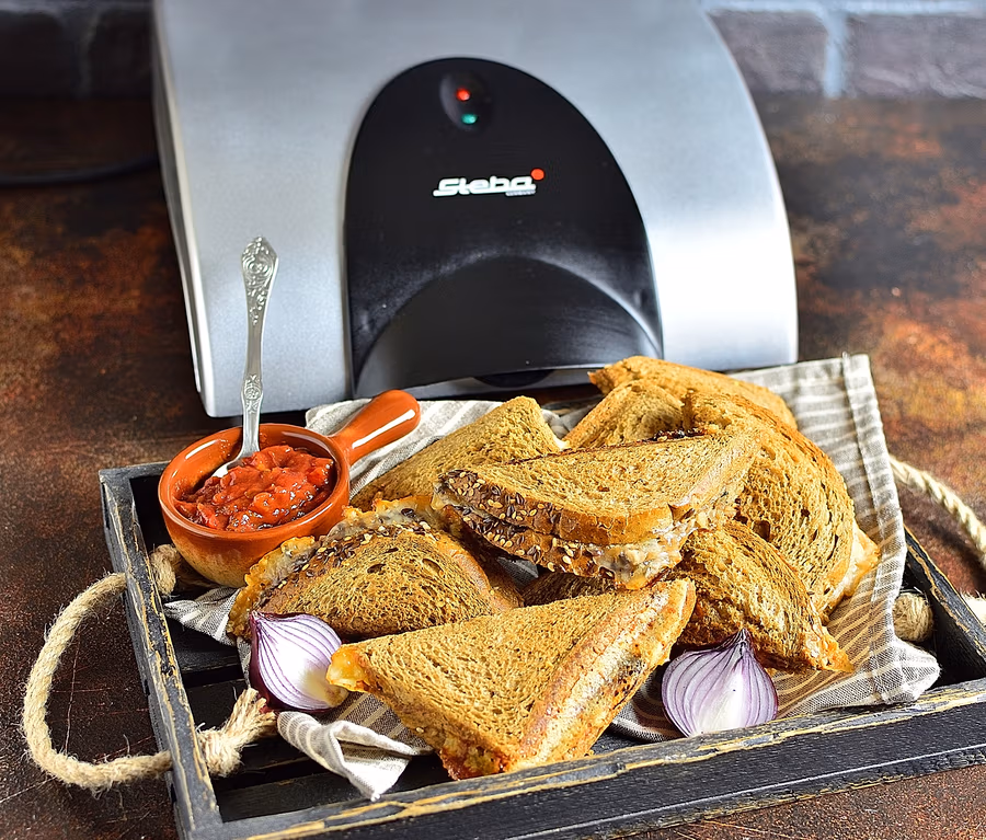 Sandwiches auf einem Tablett mit Soße und Zwiebeln neben einem Sandwichmaker Steba 4er-Sandwich-Maker »SG40«.