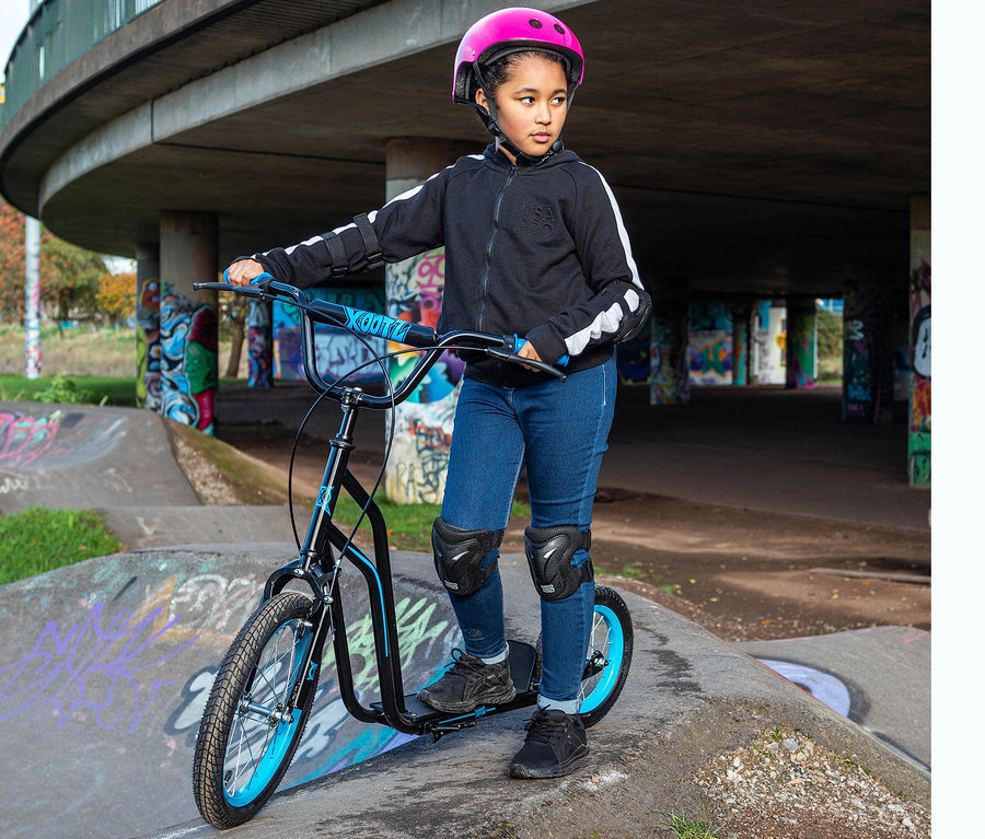 Ein junges Mädchen mit einem rosa Helm steht auf einem blauen Xootz BMX-Scooter in einem Skatepark.