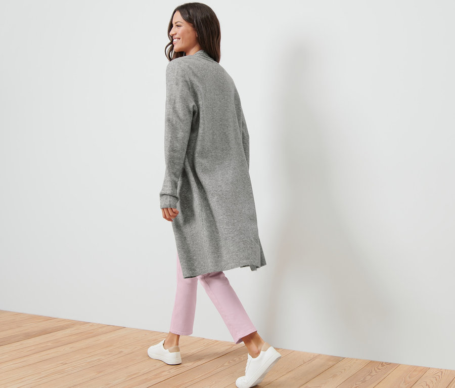 Frau geht und trägt einen offenen Longcardigan, eine rosafarbene Hose und weiße Turnschuhe.