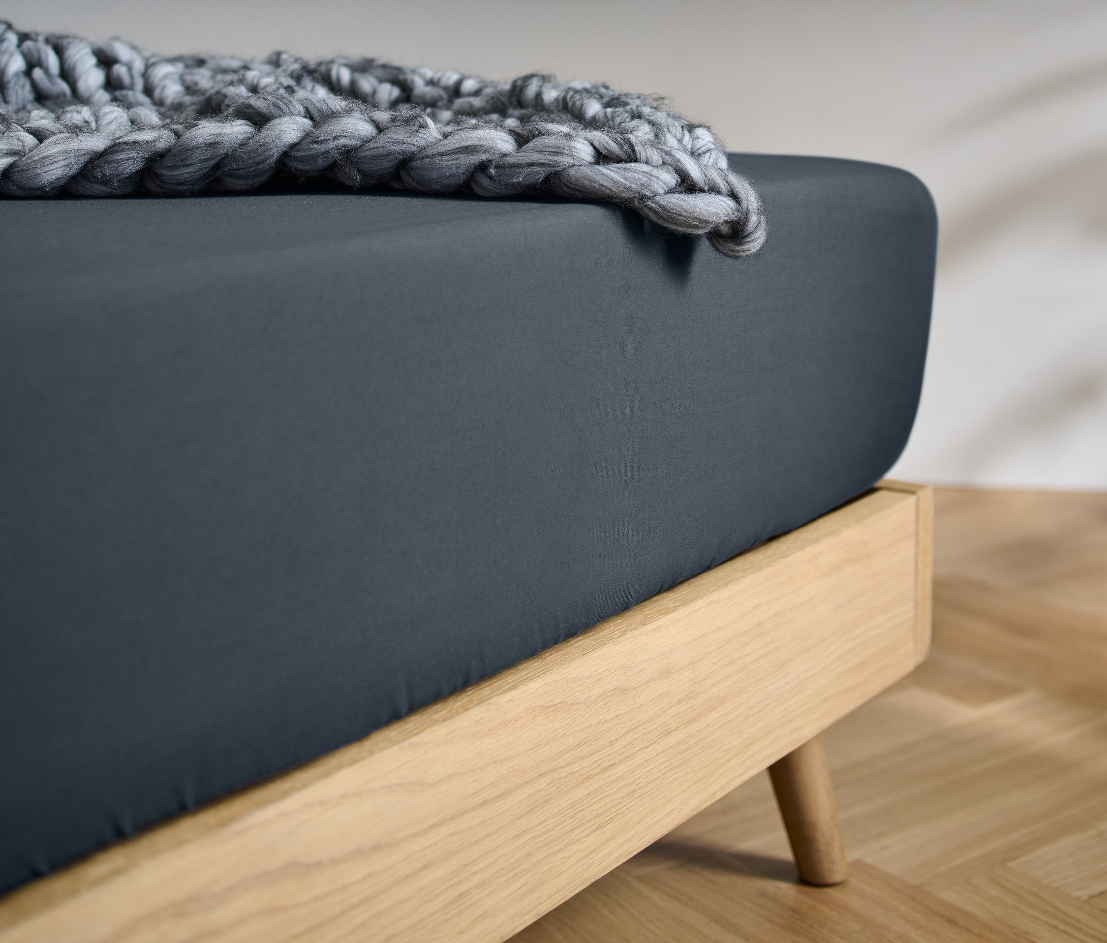 Detailansicht eines Bettes mit einem dunkelblauen Microfaser-Spannbettlaken in Normalgröße und einer gestrickten grauen Decke.