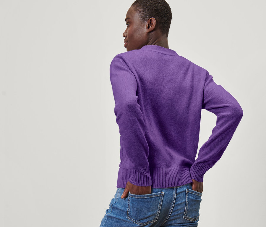 Frau posiert mit dem Rücken zur Kamera und hat die Hände in den Hosentaschen. Sie trägt eine lila Strickjacke mit Wolle.