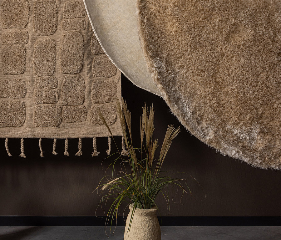 Eine beige Vase mit Pflanzen steht vor einer braunen Wand, an der zwei Teppiche hängen. Sichtbar ist der Woood Teppich »Praline« 200 cm, nougat.