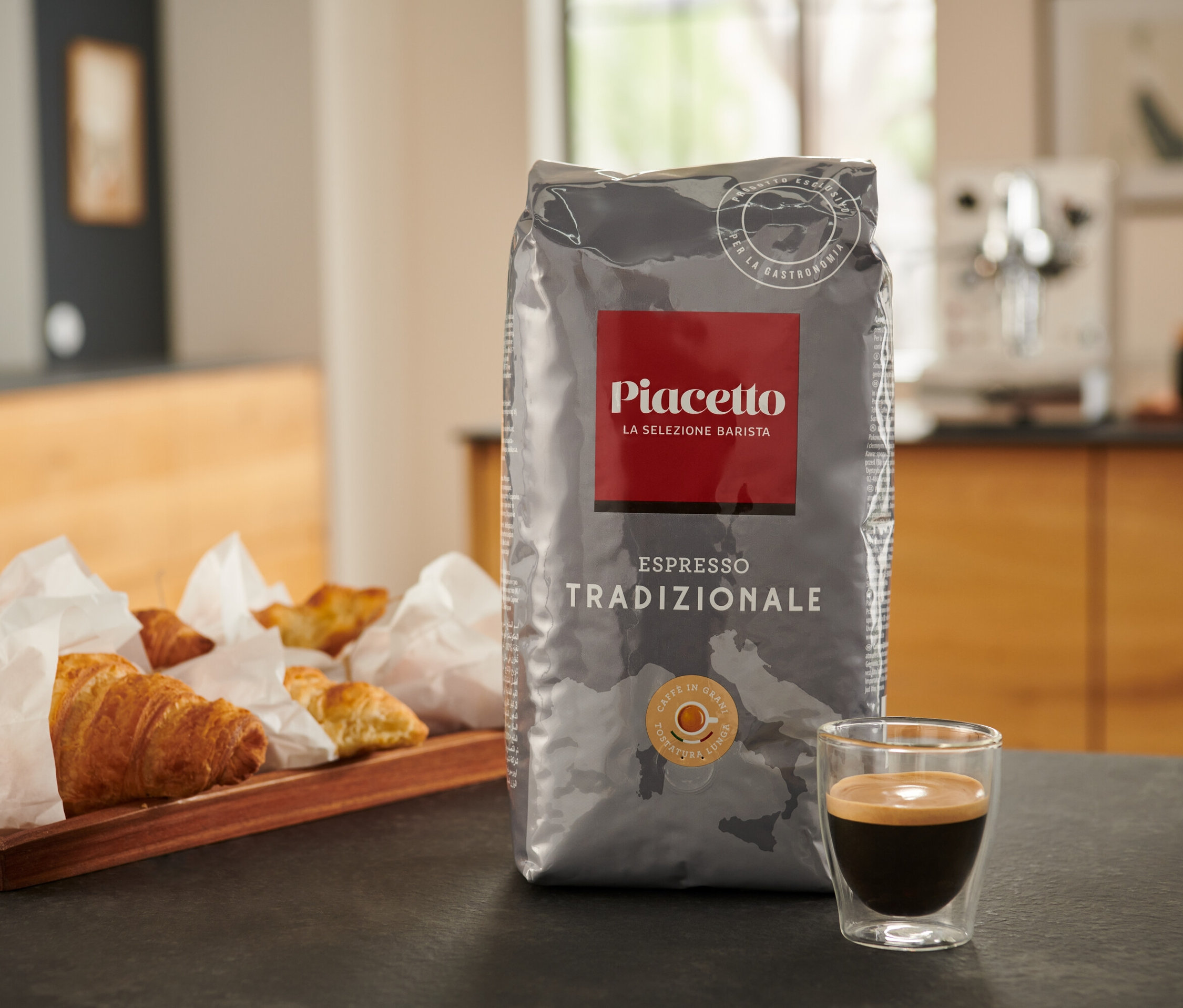 Eine Packung Piacetto Tradizionale Espresso steht auf einem Tisch neben einer Tasse Espresso und einem Tablett mit Croissants.
