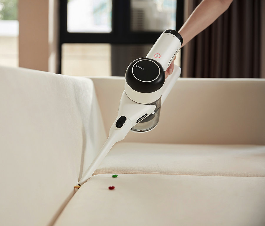 Eine Hand hält einen weißen Tineco Pure One x Essentials Akkusauger und saugt damit ein helles Sofa.