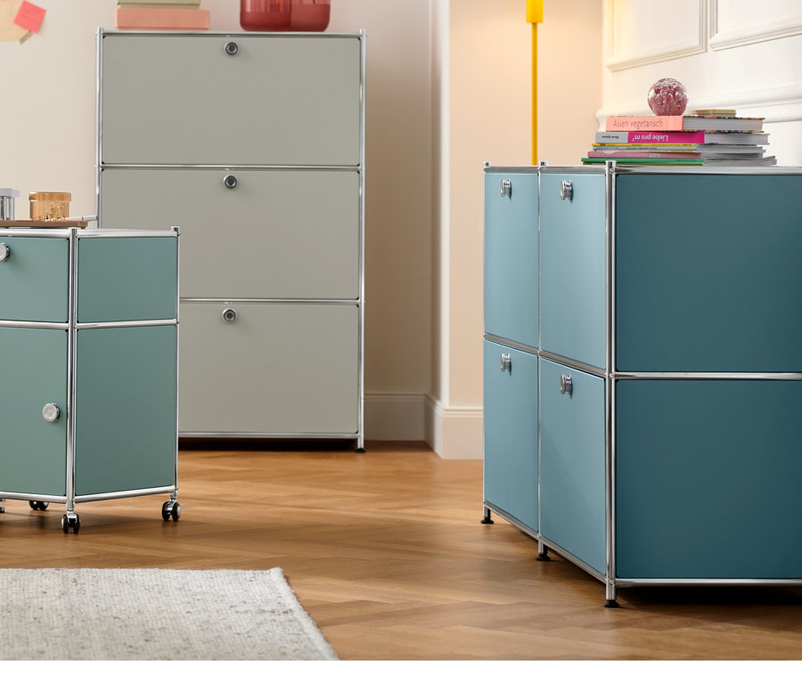 Ein weißer Schreibtisch mit einer Tischleuchte aus Metall und einem Rollcontainer »CN3« mit Schublade und Tür in Salbeigrün. Im Hintergrund eine Kommode »CN3« mit 3 Klappfächern in Lichtgrau und ein Sideboard »CN3« mit 4 Klappfächern in Basaltblau.