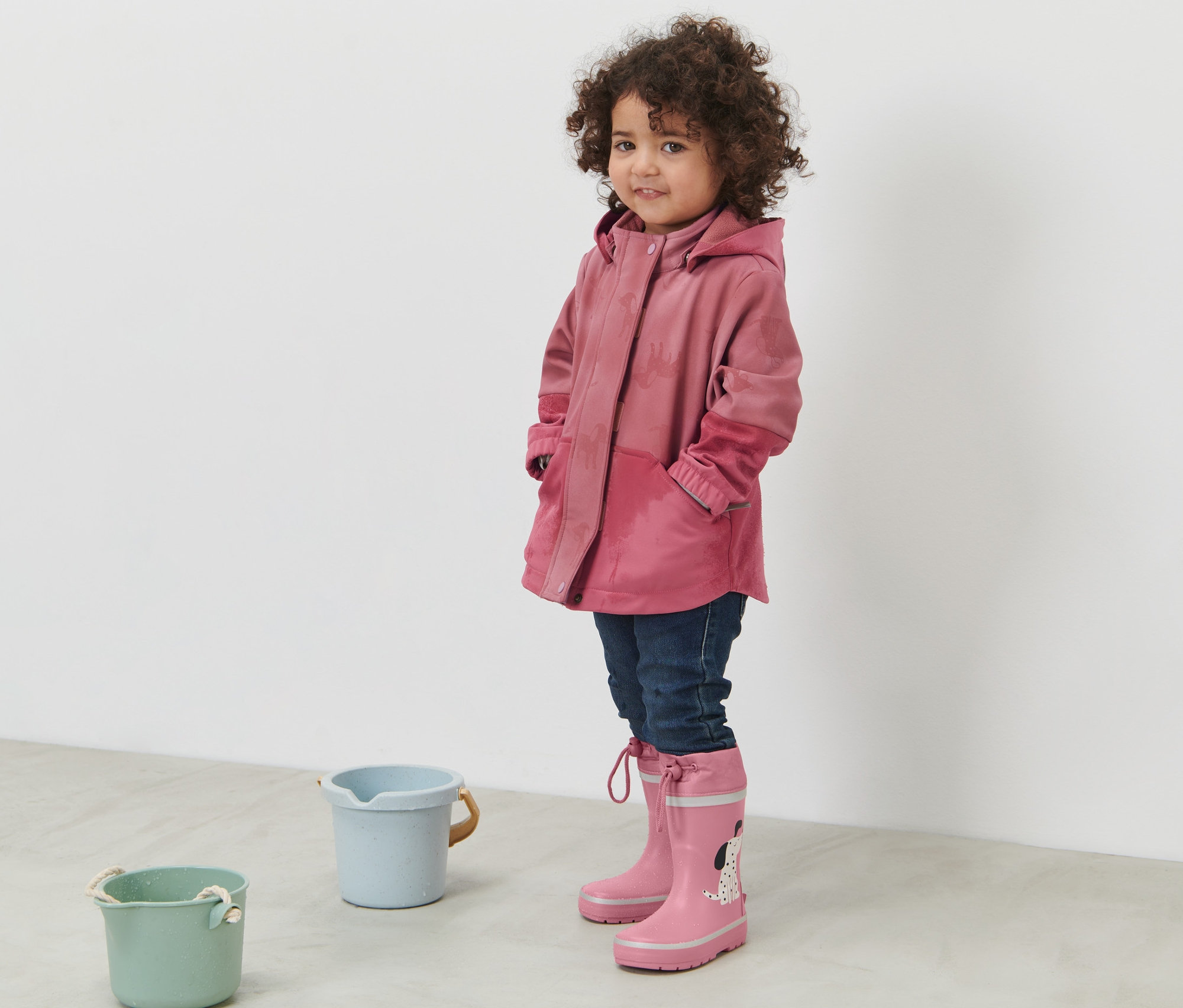 Mädchen steht und trägt eine rosa Kinder-Softshelljacke mit Farbwechsel bei Regen und rosa Gummistiefel.