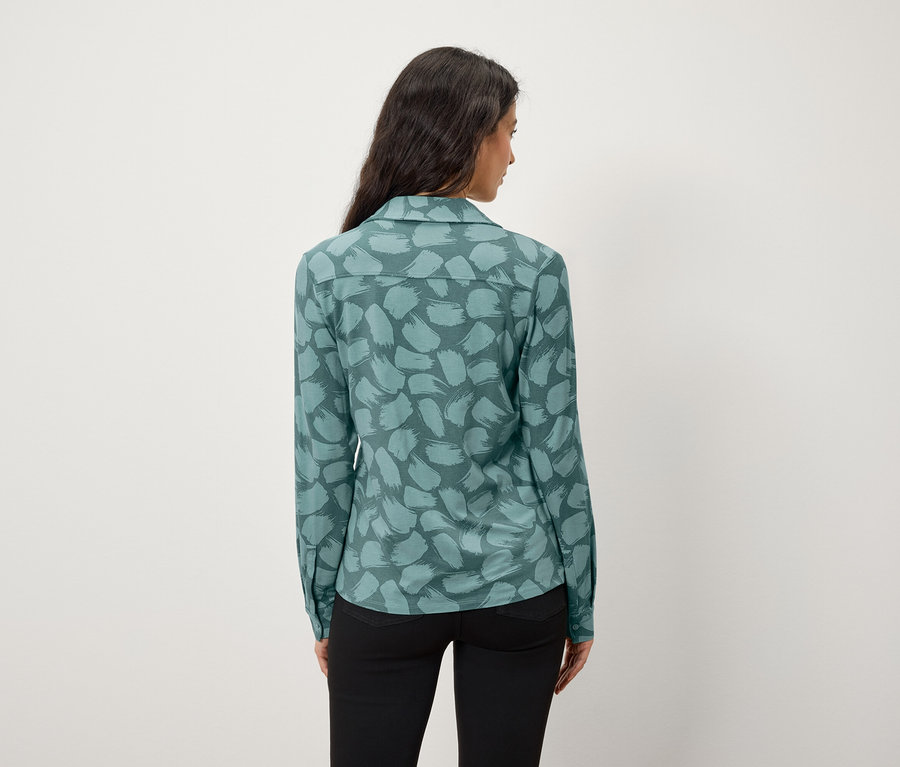 Rückansicht einer Frau mit langen, dunklen Haaren, die ein blaugrünes Hemd mit Muster und eine schwarze Hose trägt.