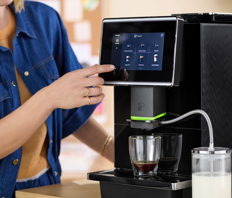Eine Frau wählt ein Getränk auf dem Touchscreen einer Kaffeemaschine aus. Zwei Glasbecher stehen auf einem Tablett, einer mit Kaffee und der andere leer. Daneben befindet sich ein Milchbehälter.
