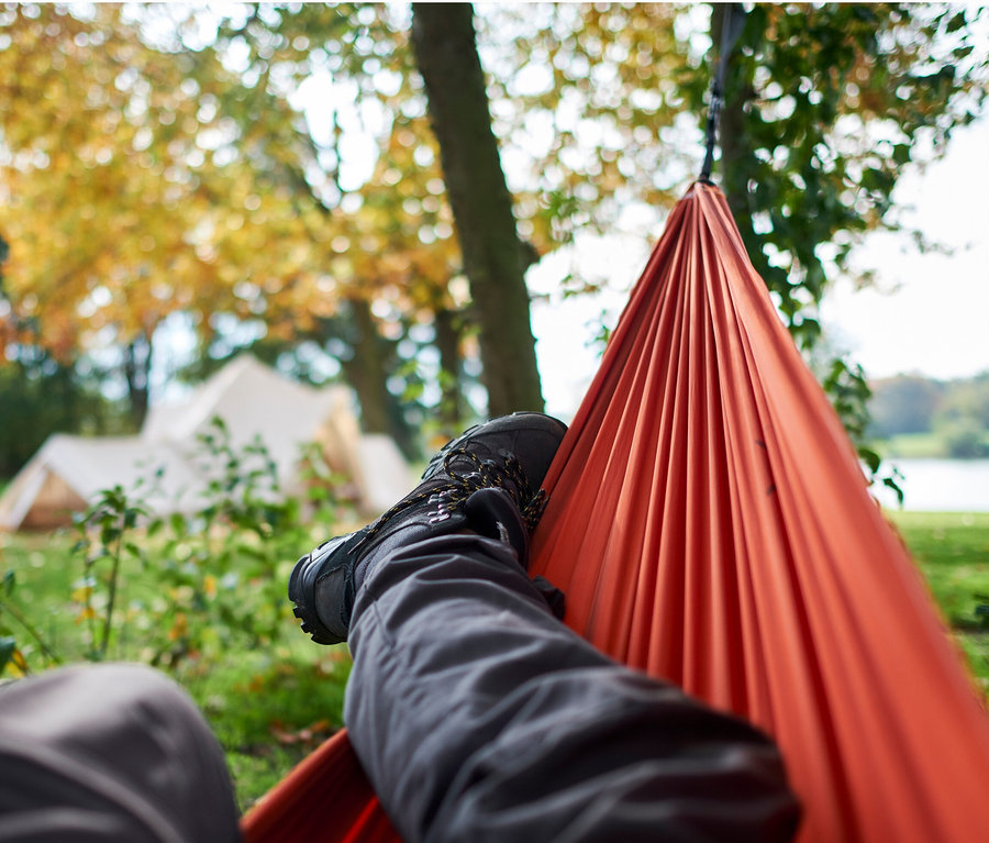 Blick aus der Perspektive einer Person, die in einer orangefarbenen Grand Canyon Bass Hammock Hängematte liegt, im Hintergrund Bäume und zwei Personen.