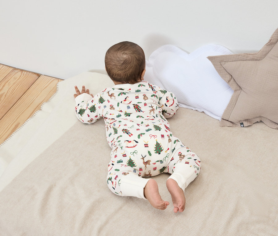 Ein Baby liegt auf dem Bauch auf einer Decke mit Weihnachtsmotiven, neben einem weißen und einem beigen Kissen.