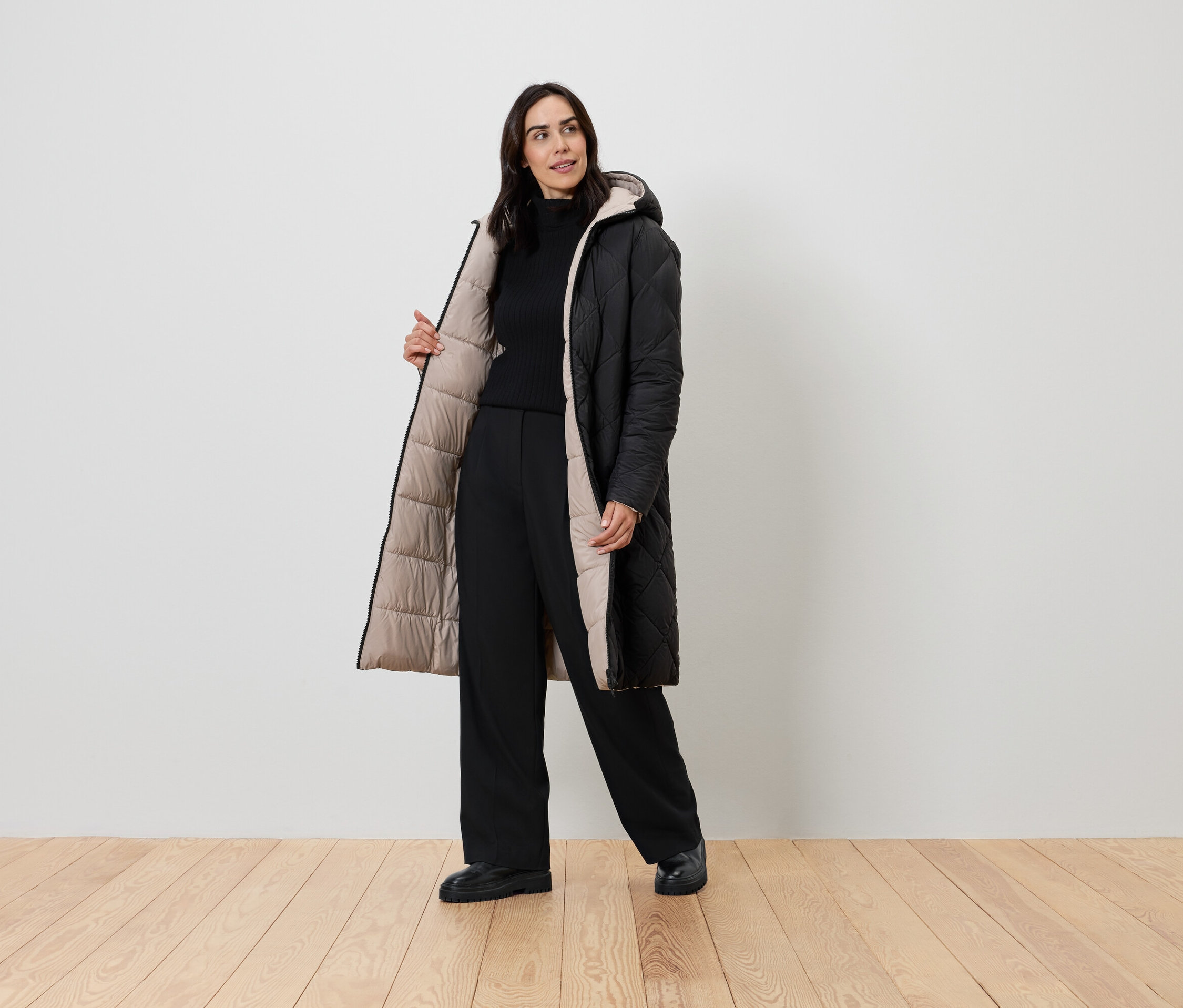 Frau posiert in einem schwarzen Mantel mit beigem Futter, einem schwarzen Rollkragenpullover und einer schwarzen Hose.