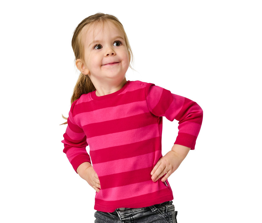 Lächelndes kleines Mädchen in einem rosa gestreiften Langarmshirt und grauen Jeans posiert mit den Händen in den Hüften.