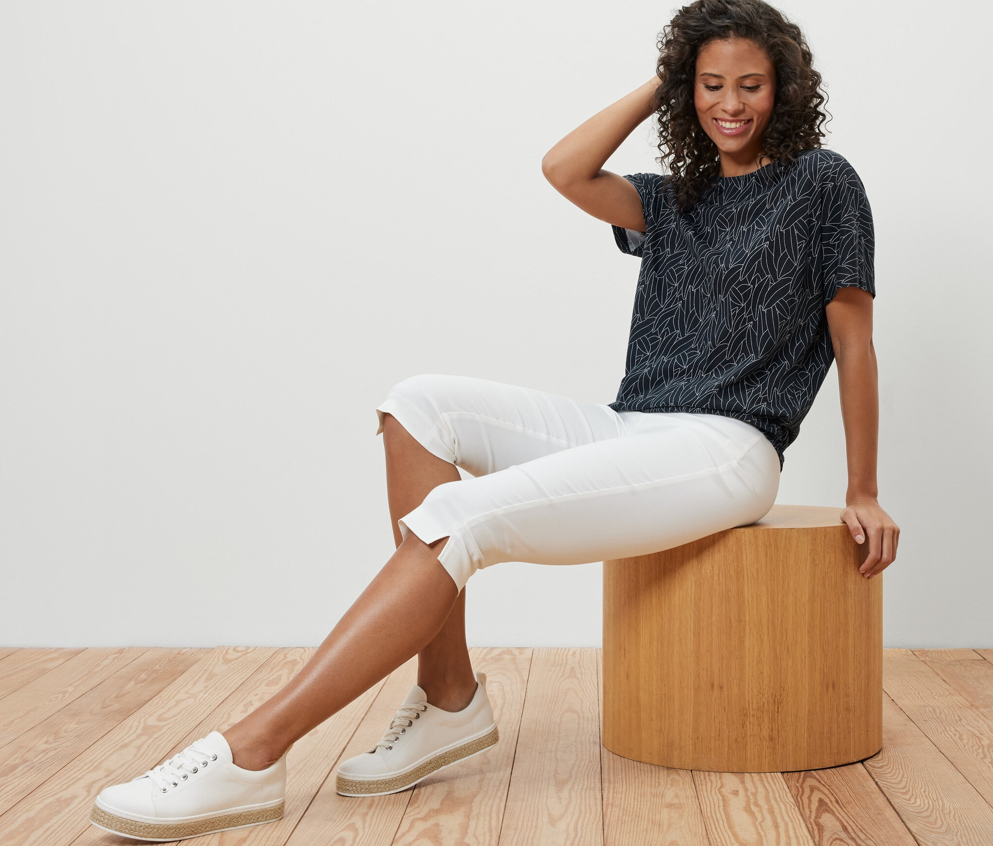 Eine Frau sitzt auf einem Hocker und trägt ein dunkelblaues, überschnittenes Blusenshirt, eine weiße Caprihose und Turnschuhe.