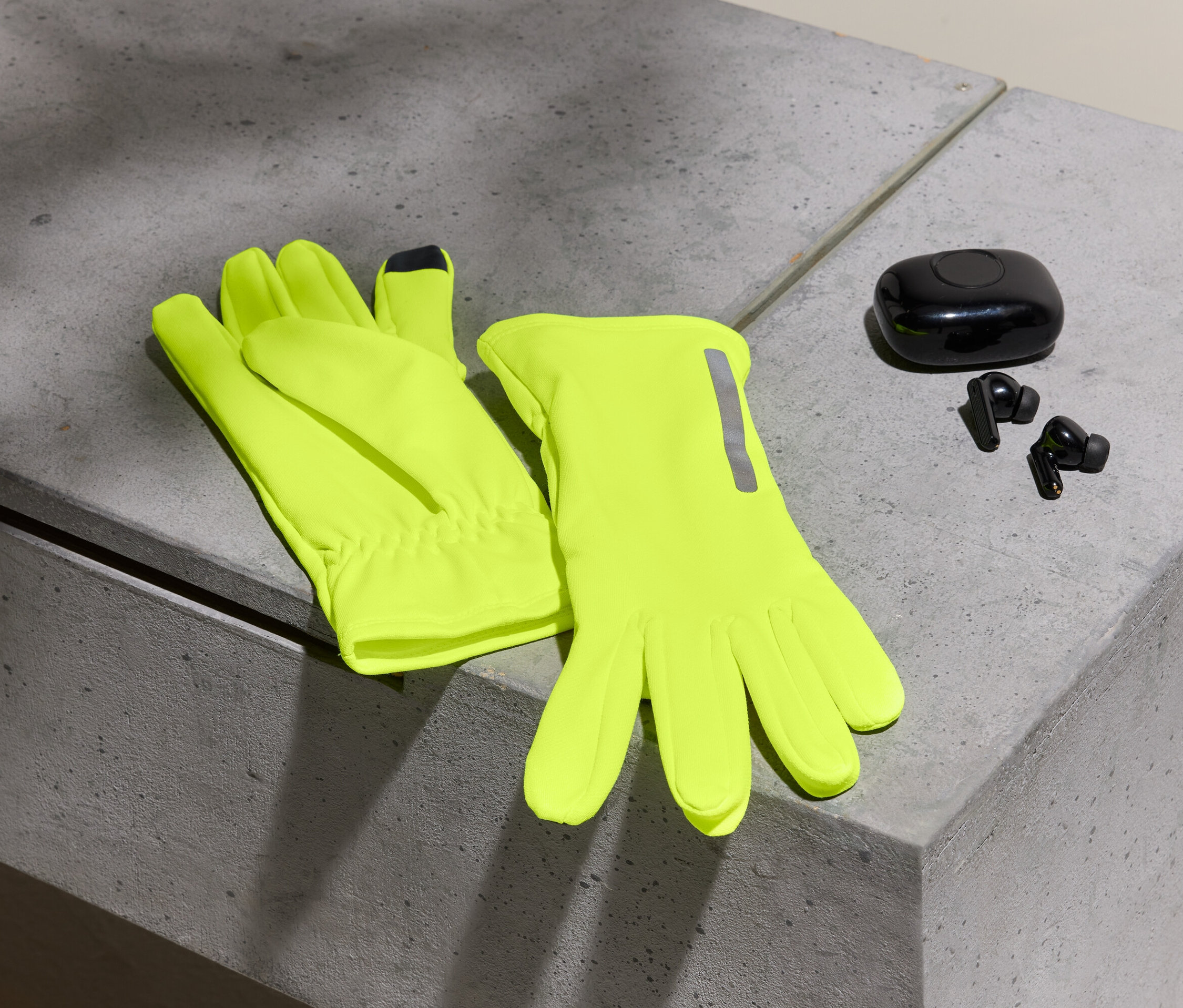 Zwei neon-gelbe Handschuhe und schwarze Ohrhörer mit Etui auf einer Betonoberfläche.