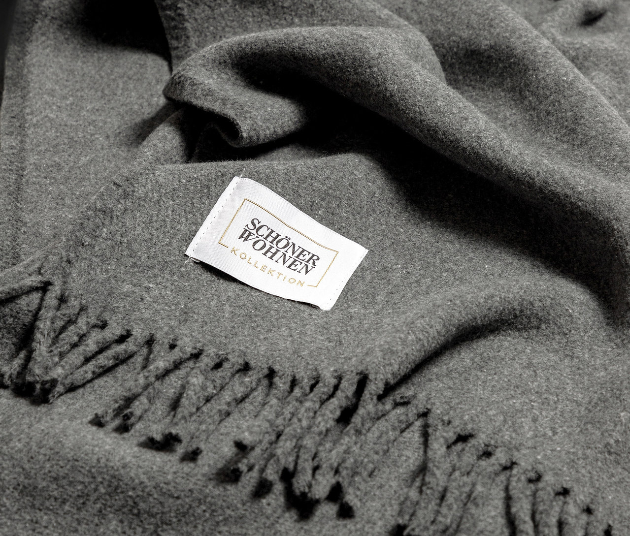 Detail einer grauen Decke mit Fransen und einem Schöner Wohnen-Etikett.