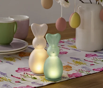 Zwei leuchtende Hasenfiguren stehen auf einer floralen Tischdecke, neben Tassen, Tellern und einem Strauß mit Ostereiern in einer Vase.