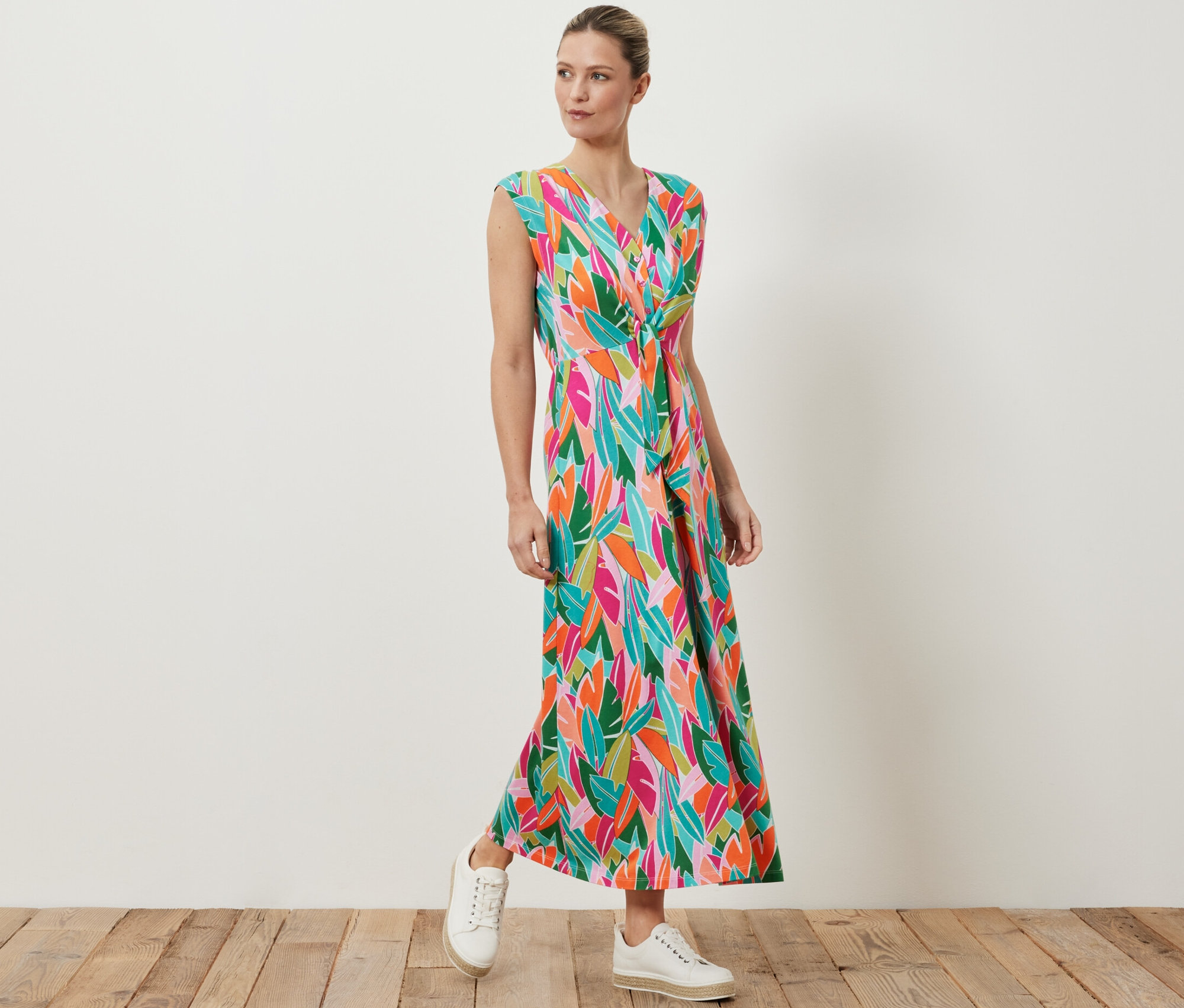 Eine Frau posiert in einem Jerseykleid mit Knoten und buntem Blattdruck, dazu trägt sie weiße Turnschuhe.