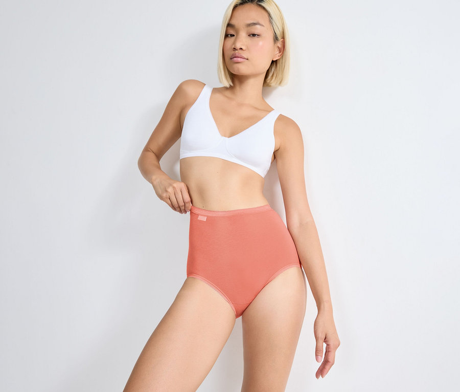 Frau in weißem BH und orangefarbenen sloggi Basic+ Maxi C3P Slips, die den Bund hält.