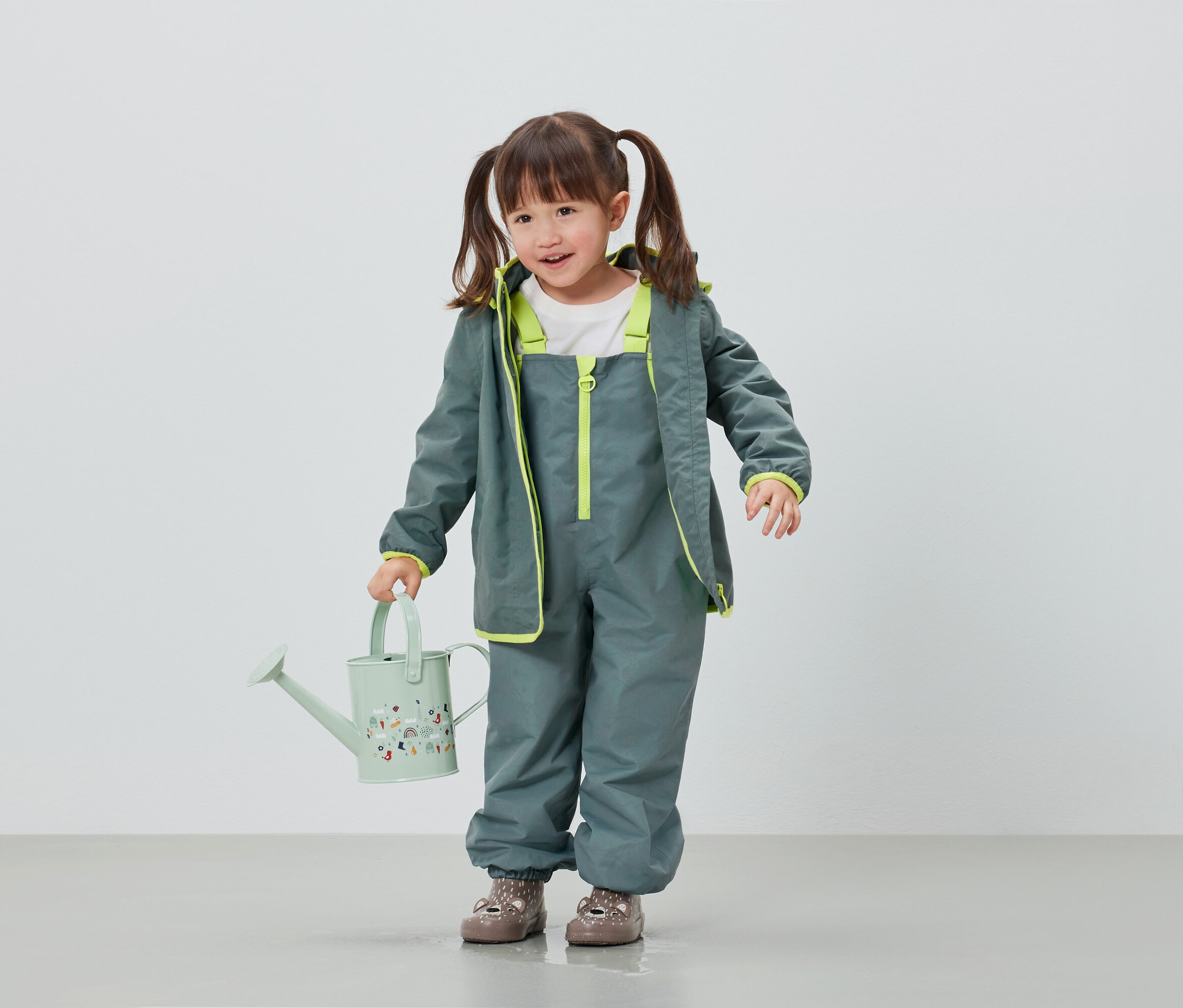 Mädchen in grauer Regenhose und Jacke hält hellgrüne Gießkanne.