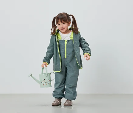 Mädchen in grauer Regenhose und Jacke hält hellgrüne Gießkanne.
