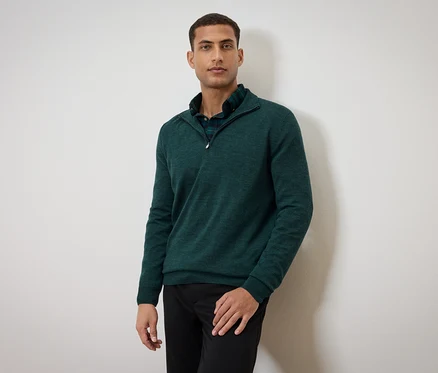 Mann posiert in grünem Pullover und schwarzen Hosen.