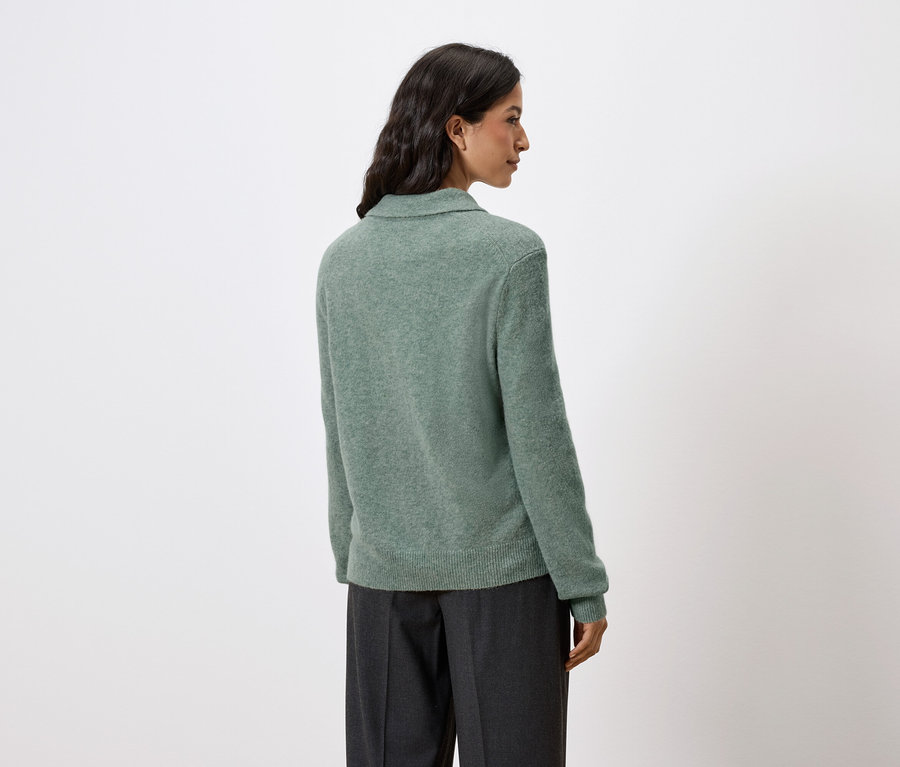 Rückansicht einer Frau mit dunklen Haaren, die einen grünen Pullover und eine dunkle Hose trägt.