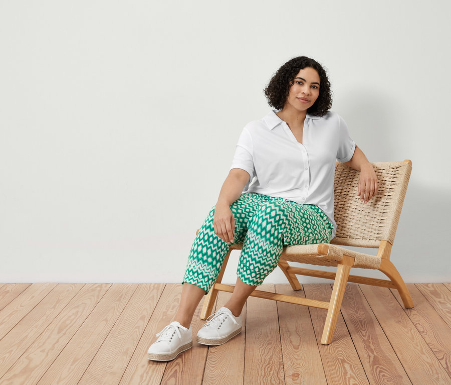 Eine Frau sitzt in einem Stuhl und trägt eine bedruckte 3/4-Jerseyhose und ein weißes Blusenshirt mit Knopfleiste.