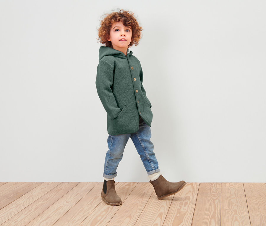 Junge mit lockigem Haar geht auf einem Holzboden und trägt einen grünen Wollmantel mit Kapuze, Jeans und braune Stiefel.