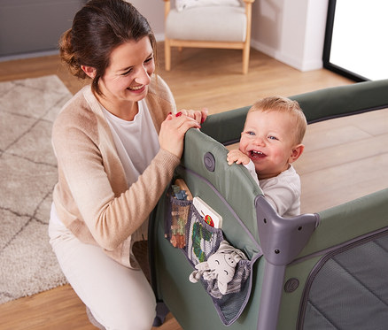 Lächelnde Frau beugt sich über ein olivgrünes Kinderbett, in dem ein lächelndes Baby steht.