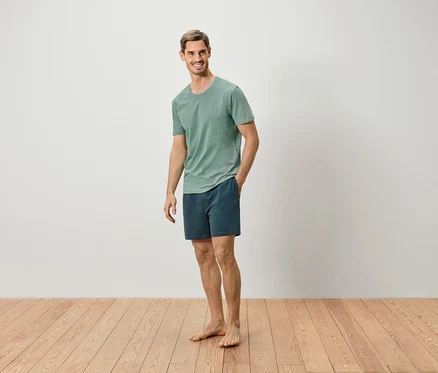 Mann steht barfuß auf Holzboden, trägt ein grünes T-Shirt und blaue Shorts.