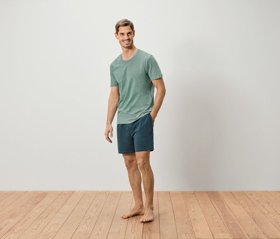 Mann steht barfuß auf Holzboden, trägt ein grünes T-Shirt und blaue Shorts.