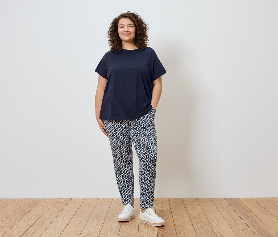 Frau steht vor einer Wand, trägt ein marineblaues T-Shirt, gemusterte Hose und weiße Schuhe.