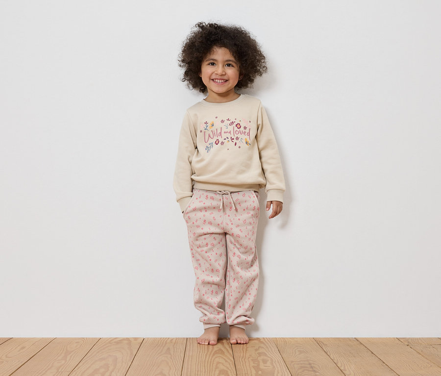 Ein lächelndes Mädchen mit lockigem Haar steht barfuß vor einer weißen Wand und trägt einen beigen Pullover mit der Aufschrift "Wild and loved" und rosafarbene Hosen.
