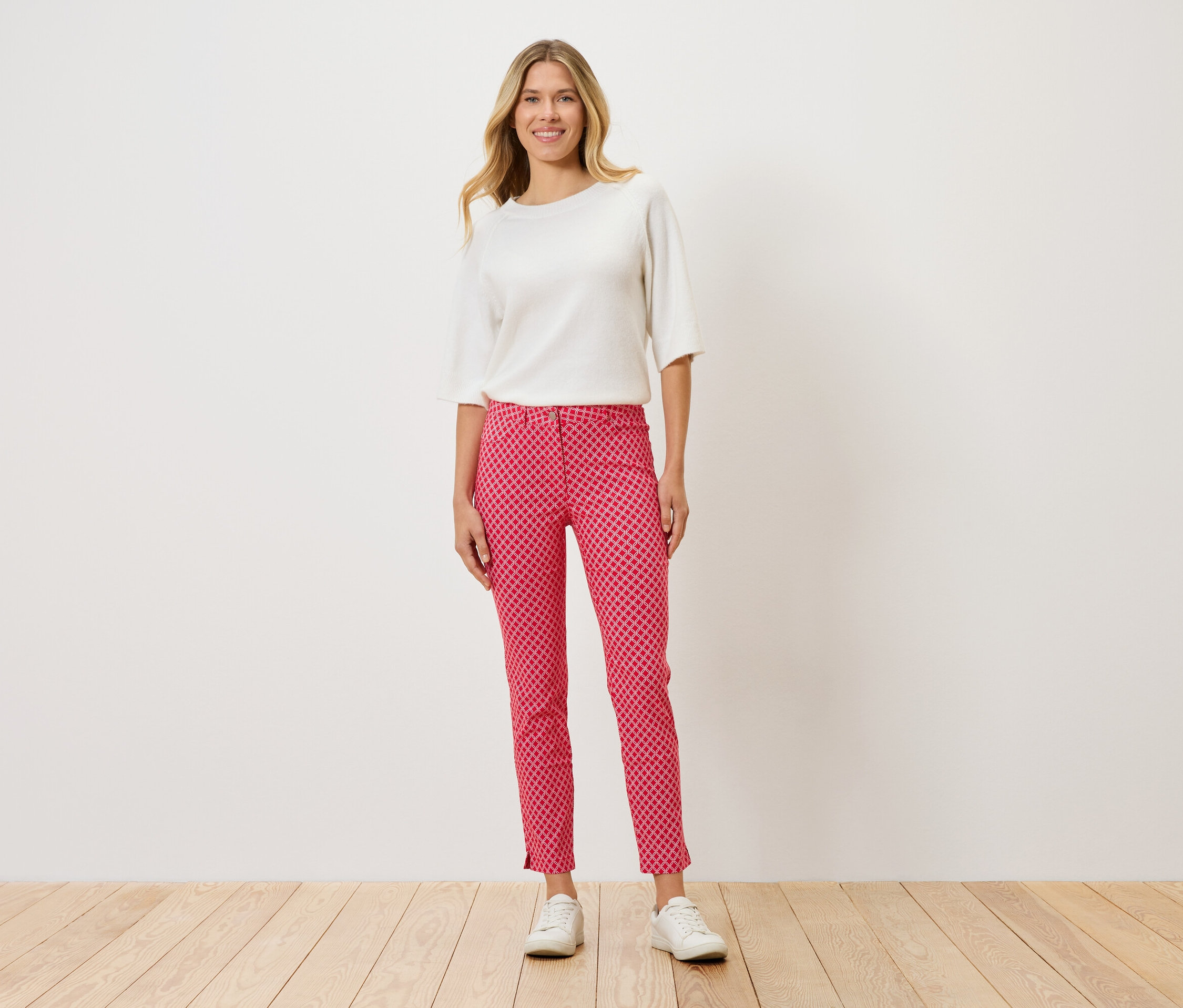 Frau steht und trägt ein weißes Oberteil, eine rote Hose mit Punktmuster und weiße Schuhe.
