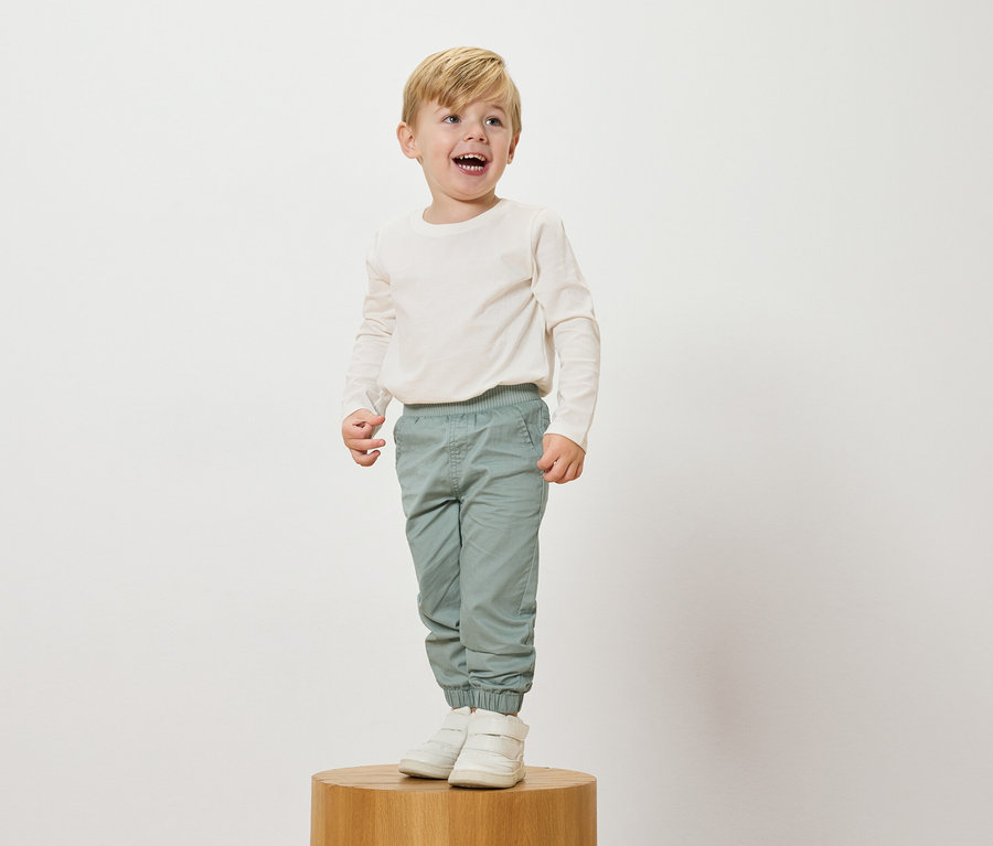 Ein lachender, blonder Junge steht auf einem Holzsockel, trägt ein langärmeliges, cremefarbenes Oberteil, hellblaue Hosen und weiße Turnschuhe.