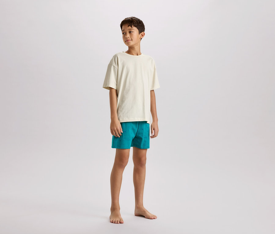 Ein barfüßiger Junge steht seitlich, trägt ein cremefarbenes T-Shirt und türkisfarbene Shorts.