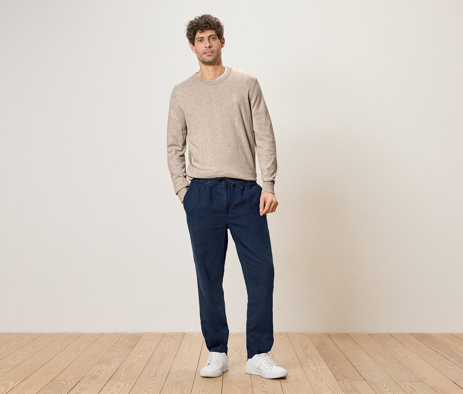Ein Mann mit lockigem Haar steht mit den Händen in den Taschen vor einer Wand, er trägt einen hellbraunen Pullover, eine dunkelblaue Hose mit Kordelzug und weiße Turnschuhe.