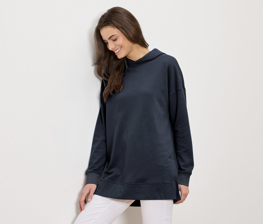 Frau mit langen braunen Haaren, die einen langen marineblauen Hoodie und weiße Hosen trägt, lächelt und lehnt an einer weißen Wand.