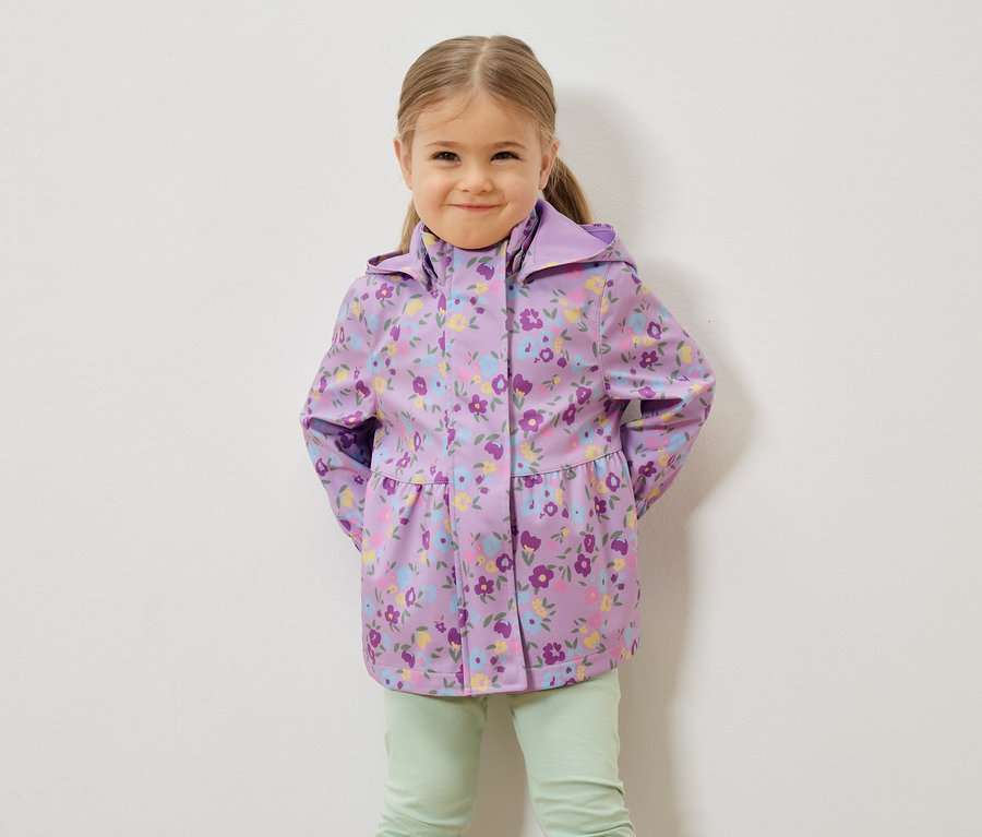 Ein junges Mädchen mit blonden Haaren trägt eine lila Regenjacke mit Blumenmuster und hellgrüne Hosen vor einer weißen Wand.