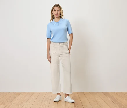 Frau steht vor einer Wand und trägt hellblaues Poloshirt, weiße Hose und weiße Schuhe.