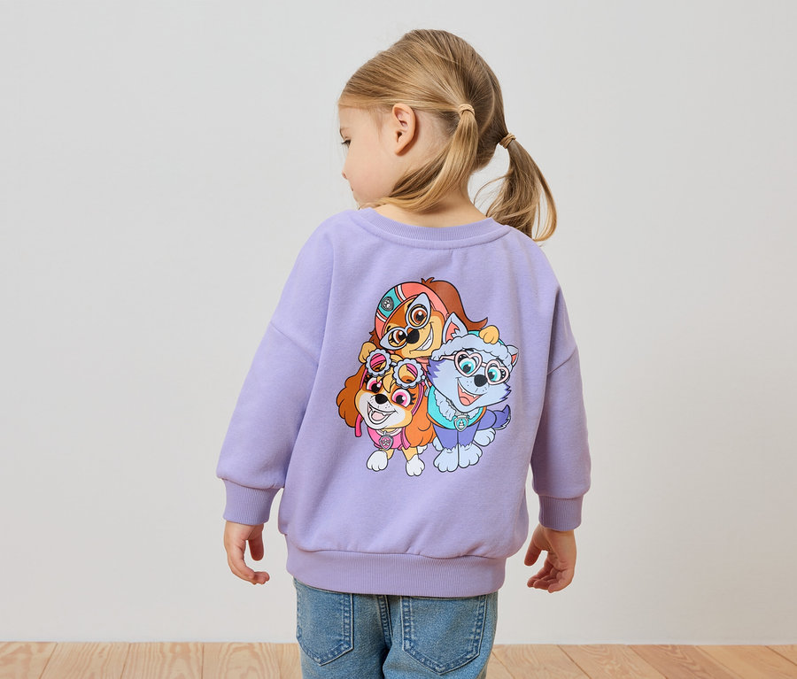 Rückansicht eines kleinen Mädchens mit blonden Haaren, das ein lila Sweatshirt mit einem Paw Patrol-Aufdruck und Jeans trägt.