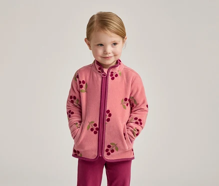 Ein kleines Mädchen mit blonden Haaren trägt eine rosa Fleecejacke mit Kirschmuster und eine pinke Hose.