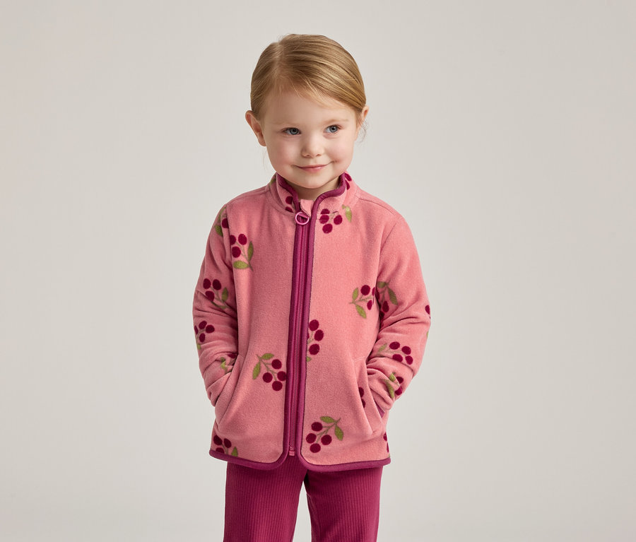 Ein kleines Mädchen mit blonden Haaren trägt eine rosa Fleecejacke mit Kirschmuster und eine pinke Hose.