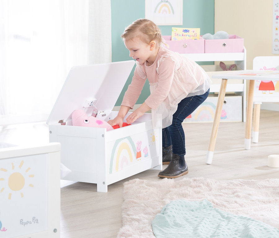 Ein Mädchen beugt sich in einem Kinderzimmer über eine offene, weiße Spielzeugkiste mit einem Regenbogen und Peppa Wutz darauf.