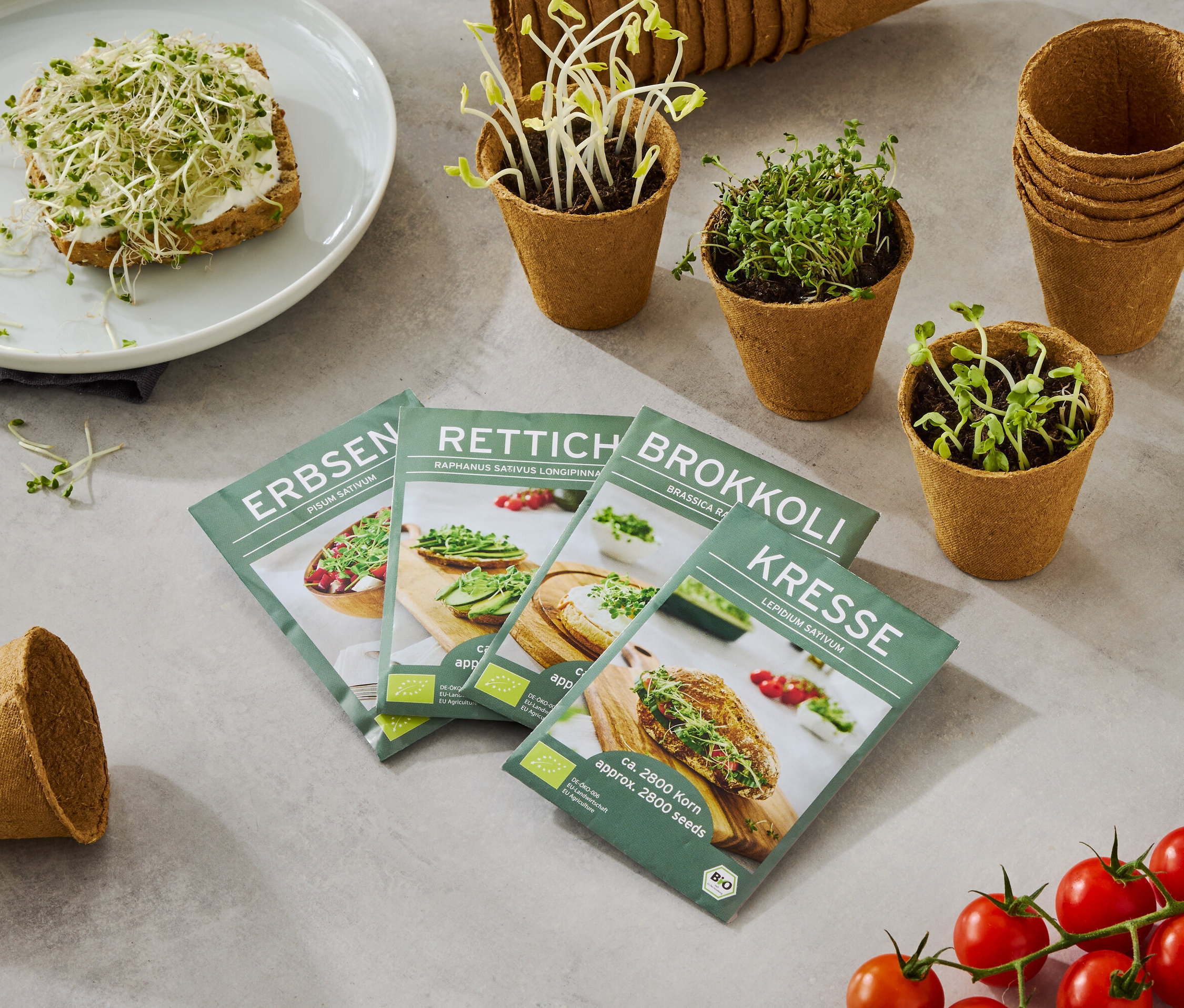 Ein Teller mit einem mit Sprossen belegten Brot, zusammen mit vier Samentüten (Erbsen, Rettich, Brokkoli, Kresse), kleinen Töpfen mit wachsenden Sprossen und einer Rispe Tomaten.